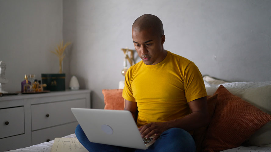 穿黄色衬衫的有色人种年轻人坐在床上用电脑