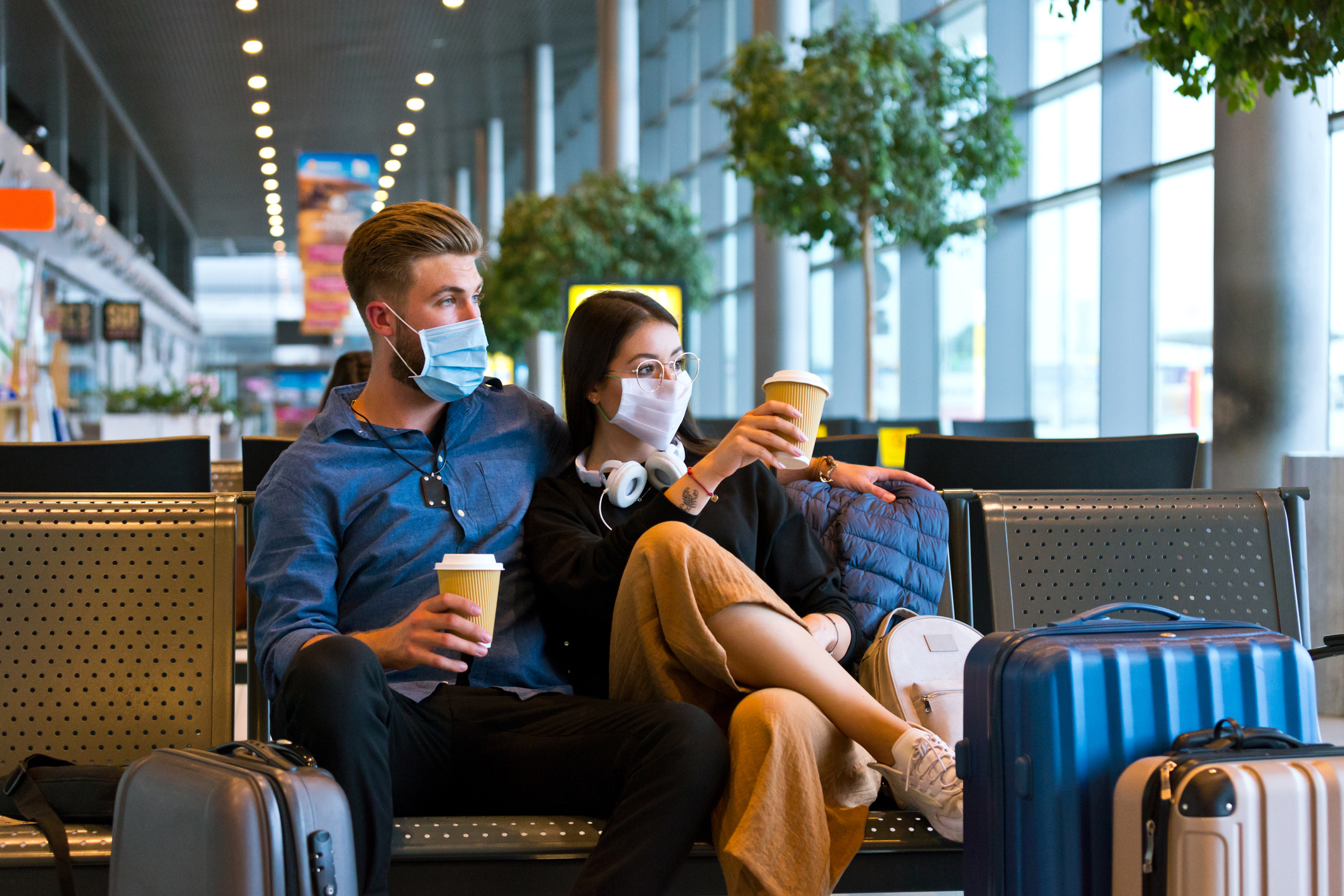 年轻男女在 COVID-19 期间乘飞机旅行，在候机区戴着 N95 口罩，拿着外卖咖啡坐在长椅上。