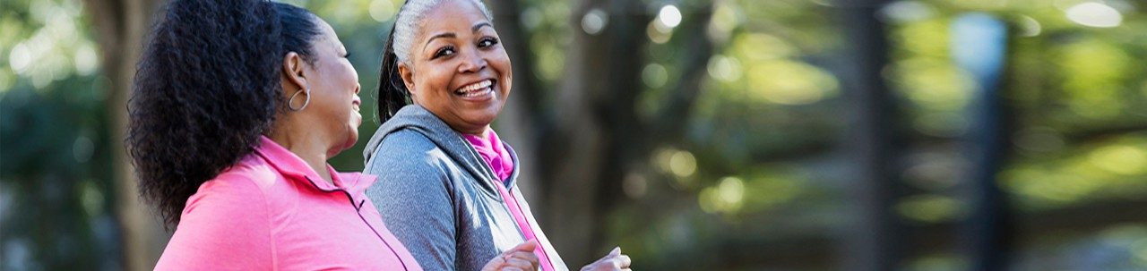 两名非裔美国女性在城市中一起运动，慢跑或快步走，谈笑风生。背景中的建筑物和树木看不清楚。穿粉色衣服的女性 60 多岁，她的朋友 50 多岁。